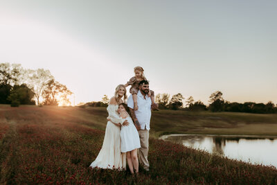 A family of four stands in a field near a pond at sunset, captured beautifully by a Warner Robins Photographer. The father holds a young child on his shoulders, the mother embraces an older child, all dressed in light clothing with trees in the background.