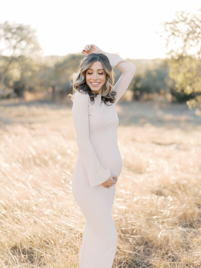 a mother to be stands in a field with beautiful golden light behind her while she smiles