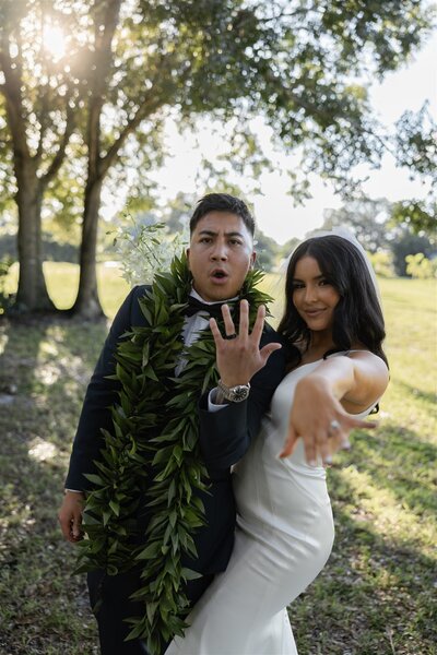 Bride and groom proudly showing off their wedding rings with playful expressions during an outdoor ceremony in Vero Beach, Florida — groom wearing a traditional Hawaiian maile lei and bride in a fitted satin gown.