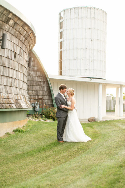 A white couple wearing wedding attire, hugging outside in front of a wood building