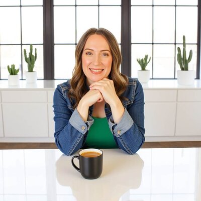 Stacey Xanthe from Xanthe Bookkeeping smiling in a modern office with warehouse windows, white cabinets, and potted cacti in the background, wearing a jean jacket and green t-shirt, seated at a glossy white table with a mug of creamy coffee.