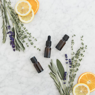 Dropper bottles of essential oils surrounded by fresh herbs and sliced citrus on marble surface
