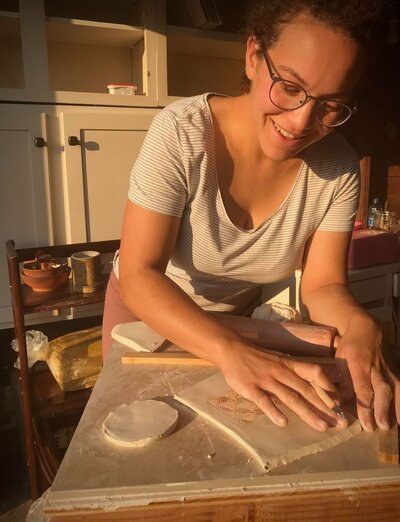 Women in golden light, wearing dark rimmed glasses and striped shirt, smiling down at her hands as she rolls out clay