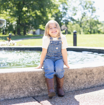 Young girl in blue jean overalls, boots, short blonde hair, white tshirt, sitting on a fountain edge in a park smiling at the camera