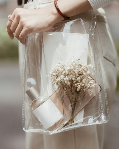 Woman carrying transparent clear bag with perfume bottle, baby's breathe flowers, white envelope, and smart phone 