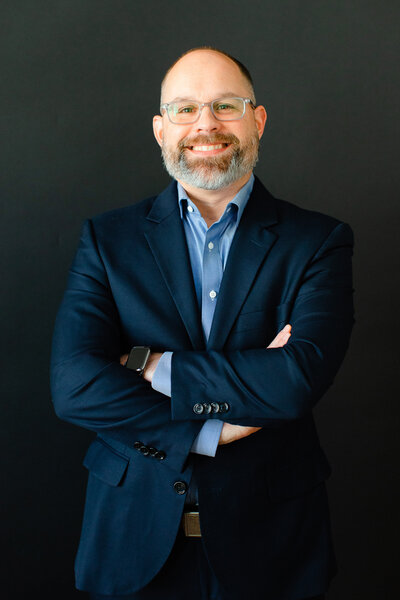 Josh Hester, founder of Storyteller Studios, stands with arms crossed, wearing a navy blue suit and a light blue dress shirt, smiling confidently against a black background.