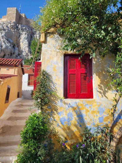 A faded yellow building with a bright red shutter surrounded by green plants and steps leading to the Acropolis in the Anafiotika district of Athens, Greece//copy