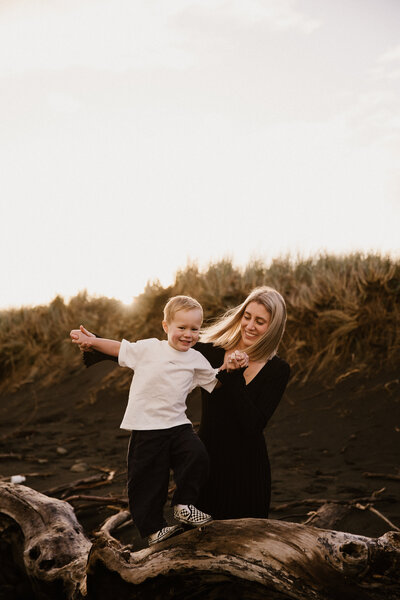 A mother helps her little boy walk along a fallen tree during a beach family photoshoot in new plymouth with aimee kelly photography