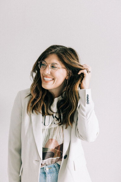 Woman in band tee and white blazer with glasses, holding her hair away from her face and smiling away from the camera