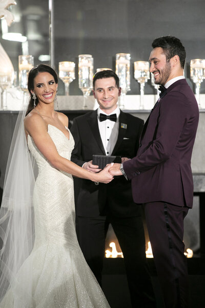 A bride in a white gown and a groom in a dark suit smile while holding hands during their wedding ceremony. A person in a tuxedo stands behind them, holding a small book. Lit candles are visible in the background.