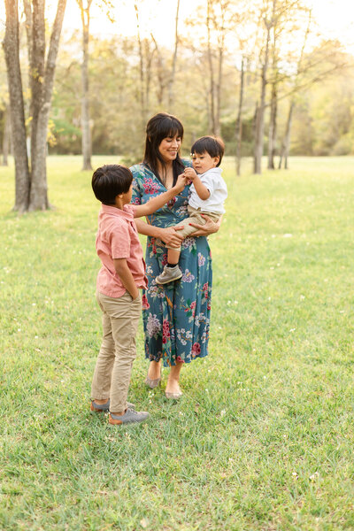 family portrait at Spring Creek Greenway Nature Center
with mom wearing a green floral dress while holding toddler in arms who is wearing a white button down with khakis and older son giving the toddler a flower