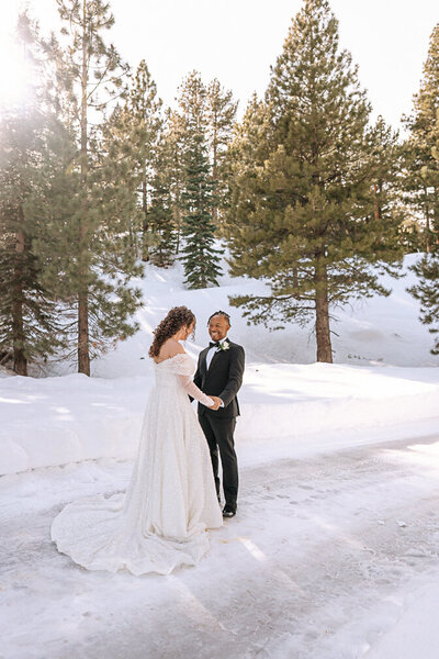 Outdoor wedding photography captures a romantic moment with a bride in an elegant white gown and a groom in a tailored olive suit. They stand before a triangular floral arch adorned with vibrant flowers, set against a serene lake backdrop. The bride's flowing dress with a long train and the groom's stylish suit add a touch of elegance to the natural setting, complete with lush greenery and golden sunset lighting.