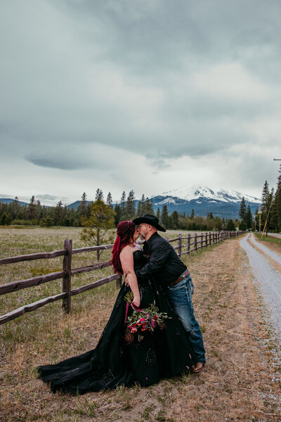 Bride in a black dress and groom in a cowboy hat share a kiss along a country fence with Mount Shasta in the background.