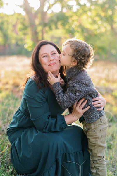 During their family photo session with Washington DC family photographer Erin Winter a mom and her young son kneel in a field during sunset. The little boy adorns his mom with a kiss on the cheek.