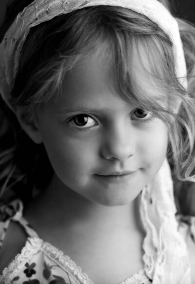Black and white portrait of a young girl with a lace headband and floral dress. She has a gentle smile and her hair is tousled softly.