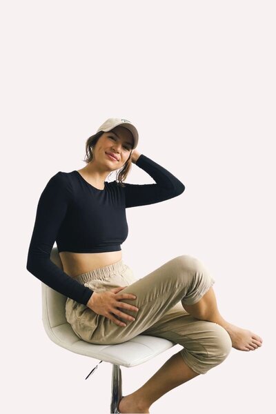 Caitlin sits comfortably on a chair, laughing with joy. She wears a black long-sleeve crop top, beige pants, and a light-colored cap against a pink background.