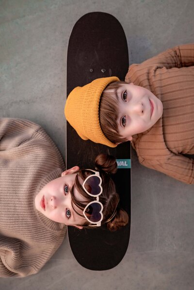 Siblings lying on the ground of a rooftop carpark resting their heads on a black skateboard