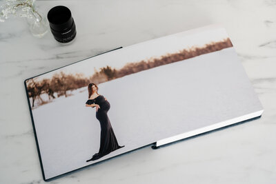 An open photo album displaying a maternity portrait of a woman in a long black dress standing alone in a snowy landscape, with barren trees in the background.