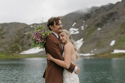 A couple embracing next to a blue lake and towering peaks. 