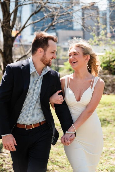 A joyful engaged couple walks hand in hand through a scenic park in Nashville, TN, with the city skyline in the background. The bride-to-be laughs as the wind tousles her hair, capturing a candid and romantic moment. This engagement session highlights the artistry of Alabama wedding photography in a vibrant urban setting.