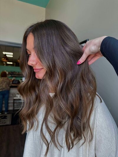 A woman with long, voluminous wavy hair, with a hand gently adjusting her hair, showcasing a full and stylish hairstyle created by 212 Salon, Spa, & Barbershop.