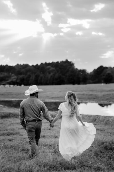 man in a cowboy hat holding hands with a woman in a white dress as they walk their a field together towards a tree line in little rock ar