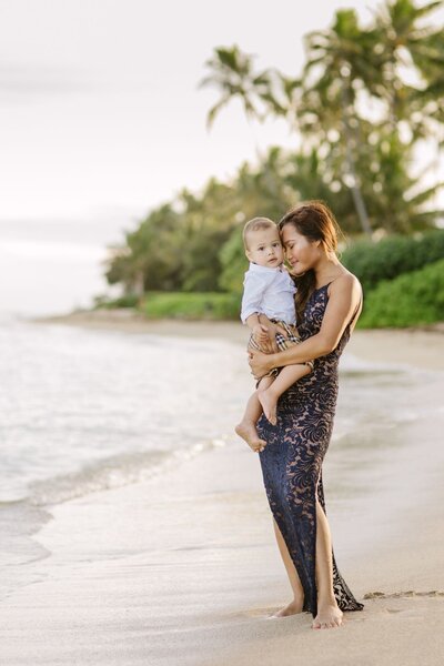A mother wearing a long black dress holds her baby boy tightly as they stand on Waikiki Beach.