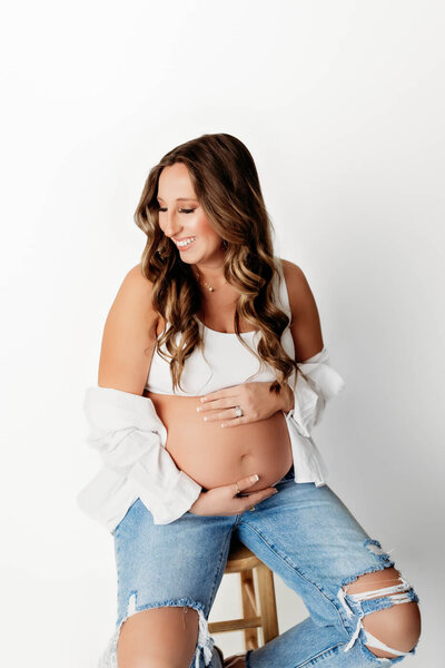 Pregnant mother sitting on a stool in front of a white backdrop. She is wearing blue jeans and a white top with her pregnant belly exposed. She is hugging her pregnant belly.