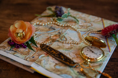 Flat lay of wedding rings, a pearl necklace, a pocket watch, and wildflowers arranged on a vintage map