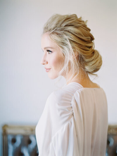 A soft bridal portrait of a blonde bride with elegant textured low bun hairstyle and natural soft glam makeup, featuring glowing skin, nude lipstick, and light blush. Captured in natural light with a minimalist background for a romantic and timeless bridal look.