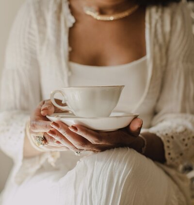 Woman dressed in white holding a teacup close to her chest, evoking sacred feminine ritual and rest.
