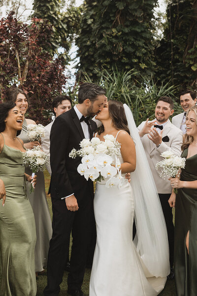 Bride and groom kiss around wedding party holding flower bouquet