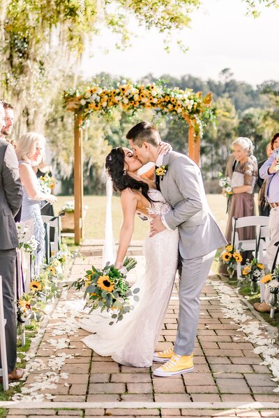 Bride and groom kiss at the alter surrounded by family and sunflowers