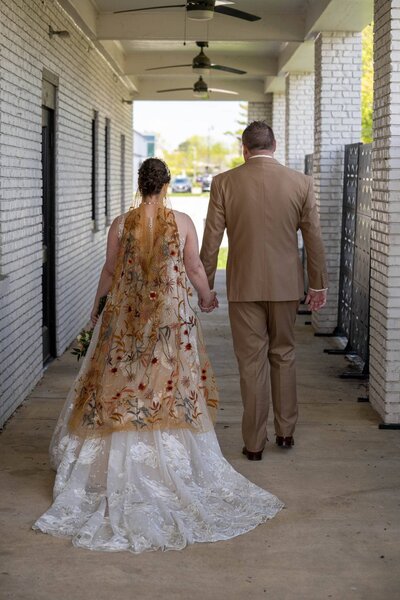 Bride and groom walking hand in hand at Bash Event Center in Carmel, Indiana, with embroidered veil and lace gown trailing behind