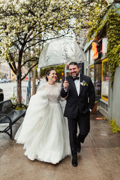 A bride in a white wedding dress and a groom in a tuxedo walk down a rainy sidewalk with a transparent umbrella above them