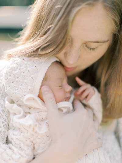 Mother's face tucked into newborn daughters face, while baby is asleep in a cream, knit bonnet
