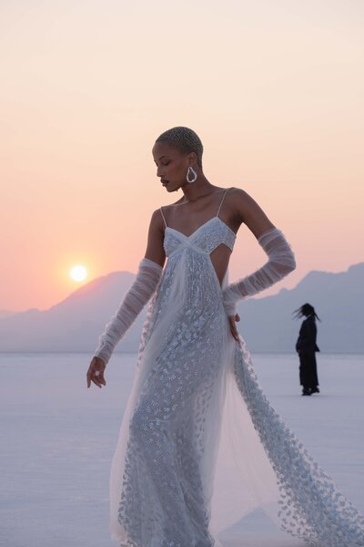 A model elegantly poses in a shimmering white gown at sunset, surrounded by a serene salt flat landscape and distant mountains.