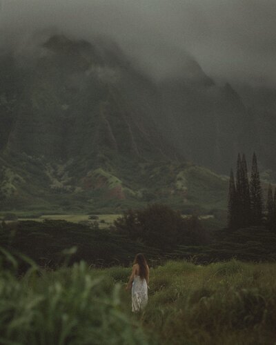 A girl with long brown hair in a white dress, standing on a lush green hill, gazing at a green mountain ahead, shrouded in mist, evoking a sense of tranquility and contemplation.