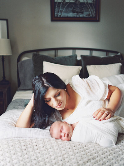 Brunette mother lies propped up in bed while placing her hand on her sleeping, newborn son taken by Photographer Little Rock Bailey Feeler