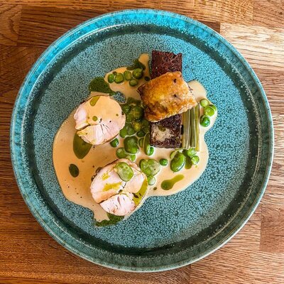 A fine dining plate featuring sliced chicken, peas, and crispy fried bread, served in a light cream sauce with garnish, presented on a blue speckled plate.