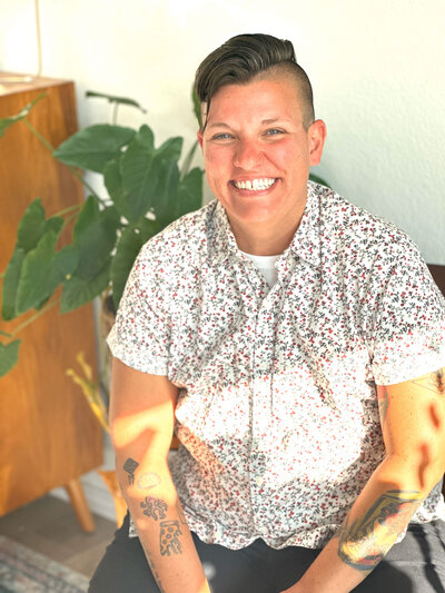 This image shows Chantille smiling toward the camera. She is sitting in a steam of sunlight, with a plant and wooden bookshelf in the background. Several small and colorful tattoos are visible on her arms.