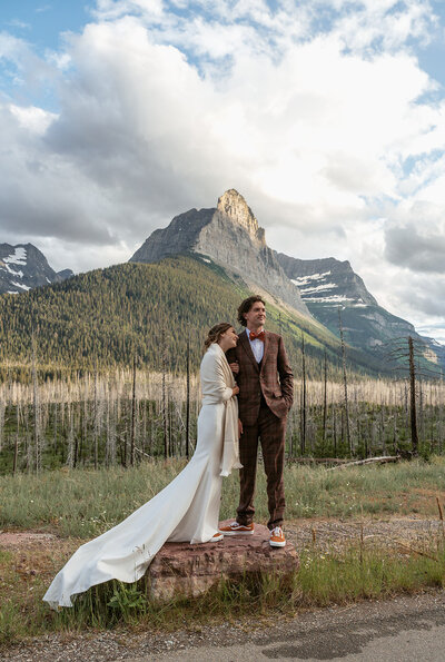 A bride and groom stand together in front of a dramatic mountain backdrop in Glacier National Park, the groom wearing a plaid suit and the bride wrapped in a shawl, captured by Sydney Breann Photography during their intimate elopement.