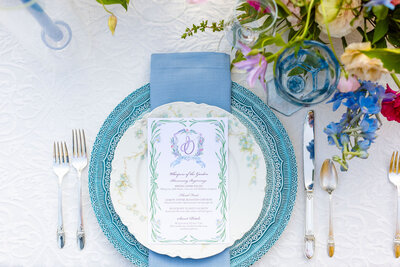 An elegant outdoor place setting featuring layered blue and white china, a pastel floral menu card, blue napkin, vintage silverware, and a vibrant floral centerpiece with blue and pink blooms on a white textured tablecloth.