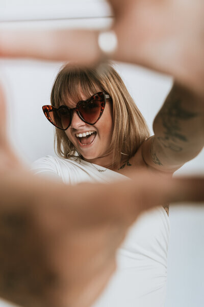 Laura Termeau, avec des lunettes en forme de cœur, visage encadré par ses mains devant un fond neutre.