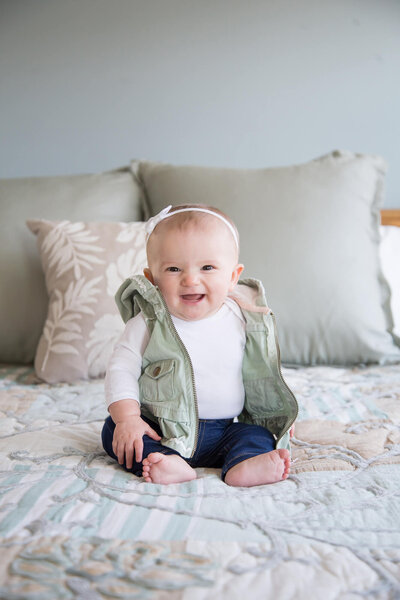 Portrait of a happy baby girl sitting up on a bed to capture the moment before it's gone. by portrait photographer, Jessica Bowles