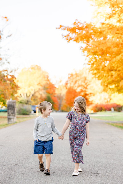 Brother and sister walk down the street holding hands and smiling at each other.