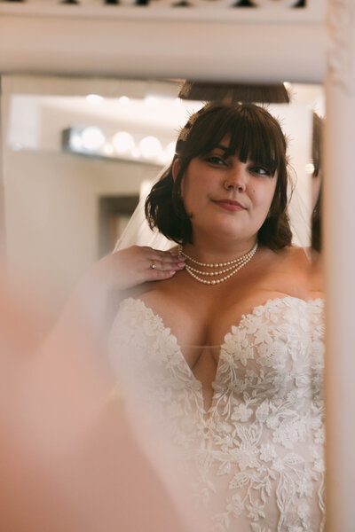 Bride admires her pearls and wedding dress in a mirror.
