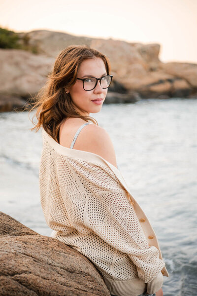A highschool senior leans against a rock at Wingaersheek Beach and stares at the camera as wind gently blows her hair.