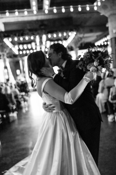 A bride and groom share a romantic kiss on the dance floor during their wedding reception. The bride wears a classic ball gown with elegant gloves, while the groom is dressed in a black suit. 
