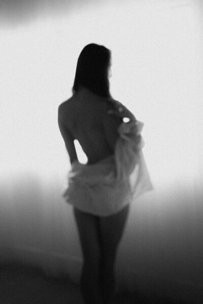 Soft, blurred silhouette of a woman standing near a bright window, wearing a loose shirt during a light-filled boudoir photography session in Phoenix.